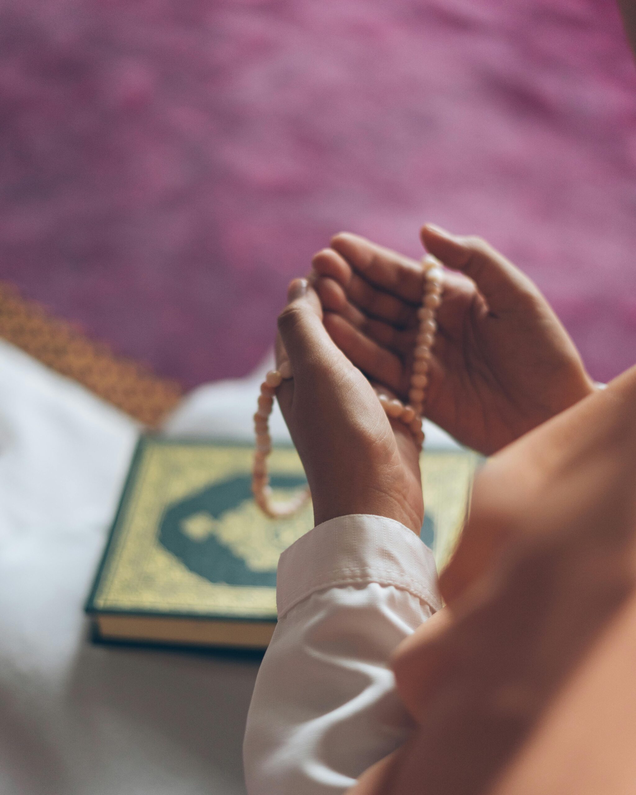 Hands holding prayer beads over an open Quran, symbolizing reflection and spirituality in a serene indoor setting.